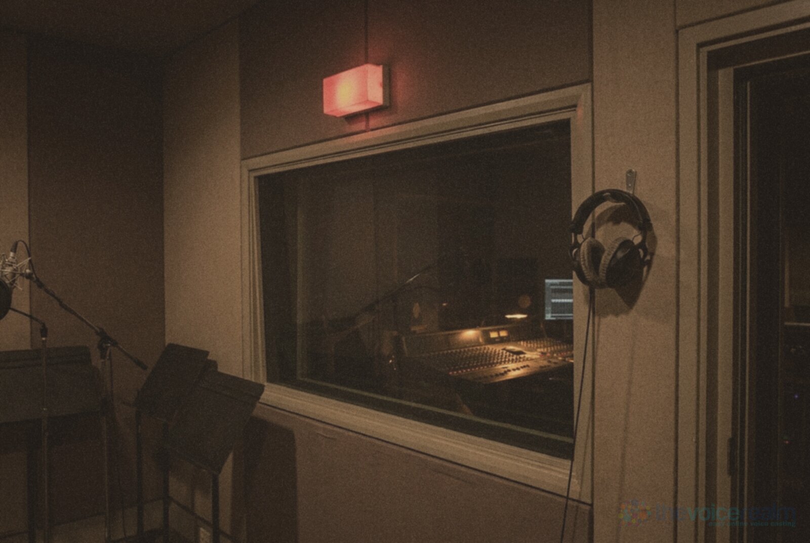 The view from inside an empty recording studio towards the control room window, a red recording light glowing above it, a mixing desk lit behind the glass and headphones hanging on a hook © The Voice Realm