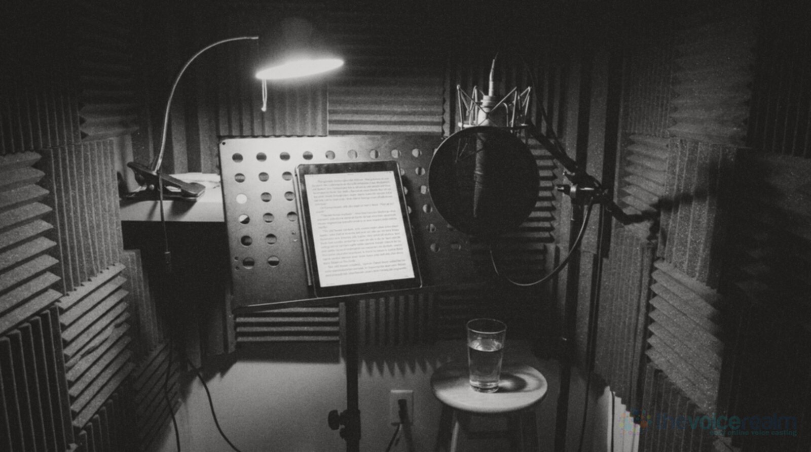 Black and white photo of a small foam-lined home booth at night, a tablet on a music stand showing a page of a novel, a microphone with a pop filter, a desk lamp and a glass of water on a stool © The Voice Realm
