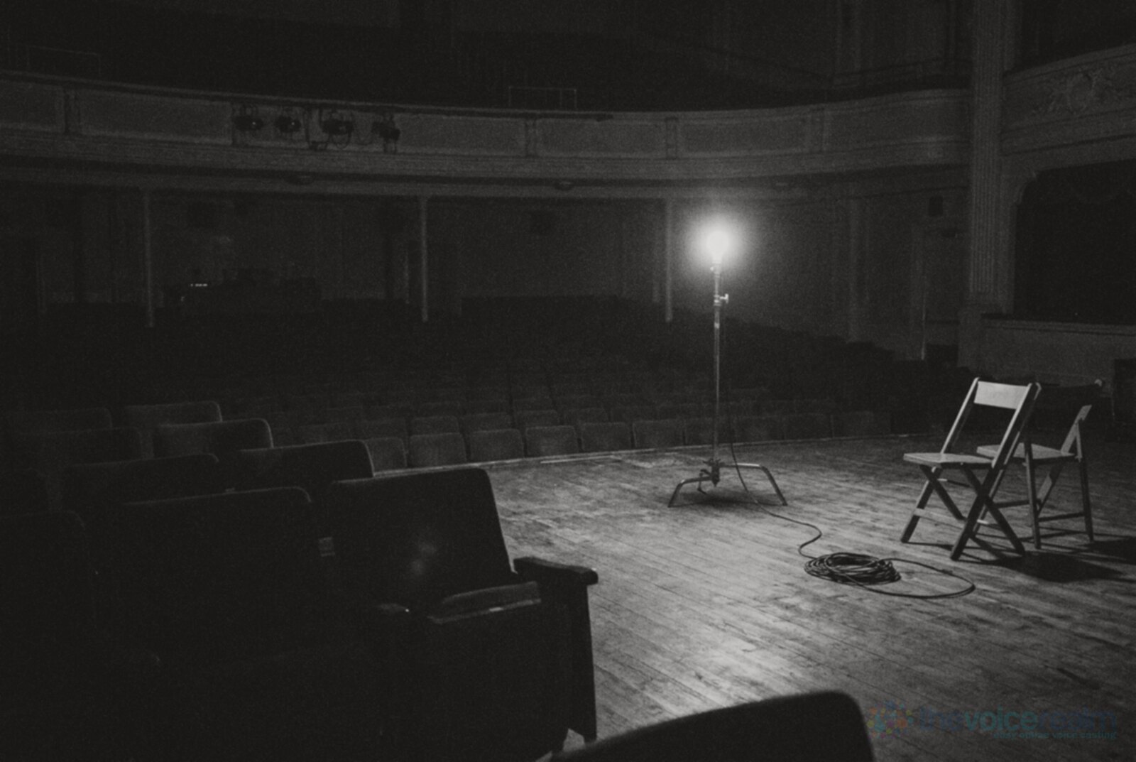 Black and white photo of an empty theatre stage at night lit by a single bare ghost light on a stand, two folding chairs on the boards and rows of empty seats in the dark © The Voice Realm