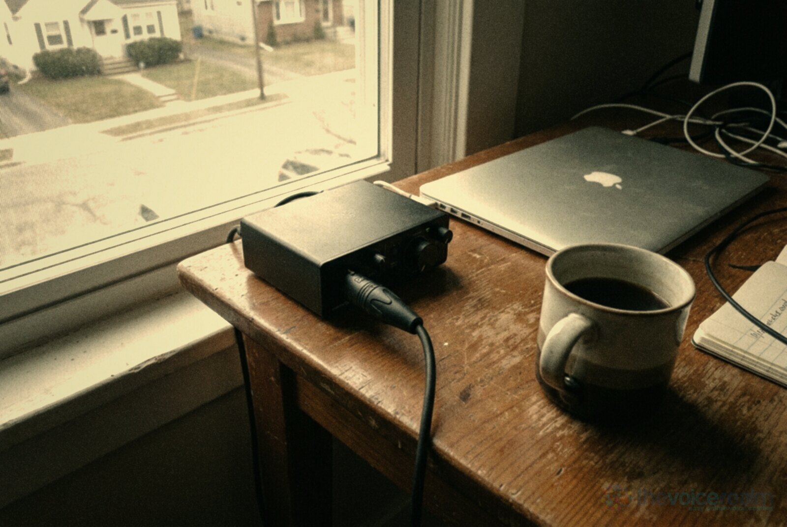 A small black audio interface on a wooden desk with an XLR cable plugged into its front input, a closed laptop and a mug of tea beside a window, in faded warm film colours © The Voice Realm
