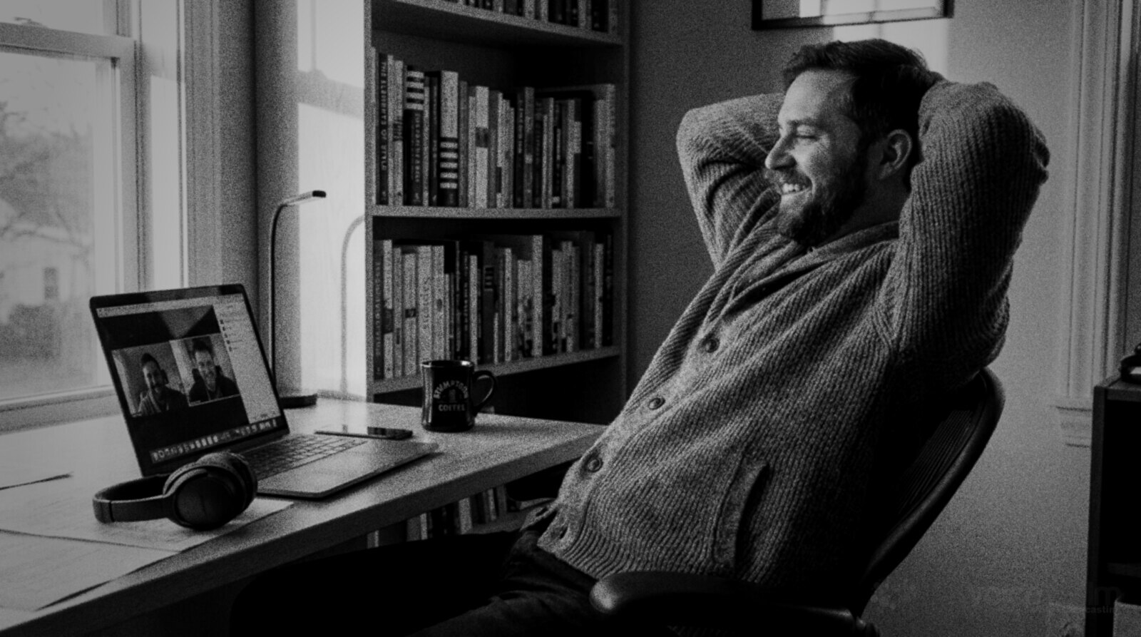 A man in a cardigan leans back in his desk chair with his hands behind his head, smiling at a video call on his laptop in a sunny home office lined with books, black and white photograph © The Voice Realm