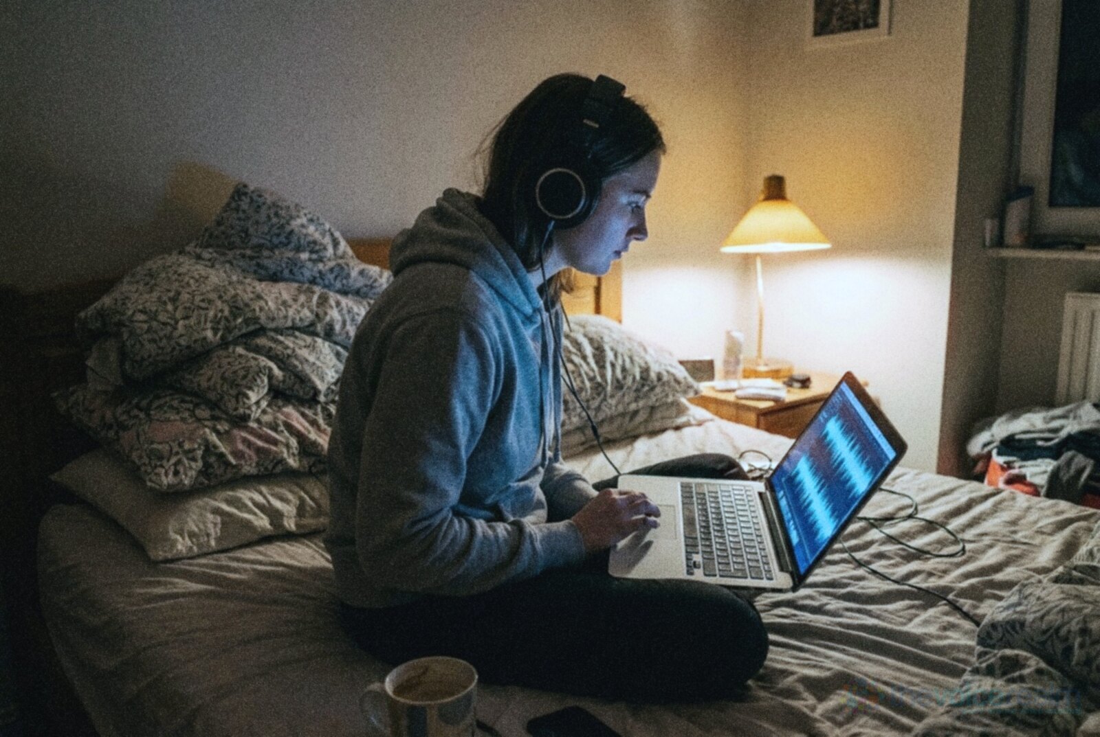 A young woman in closed-back headphones sits cross-legged on her bed editing a waveform on a laptop late at night, a duvet and pillows piled behind her and a bedside lamp glowing, in cool grainy night film colours © The Voice Realm