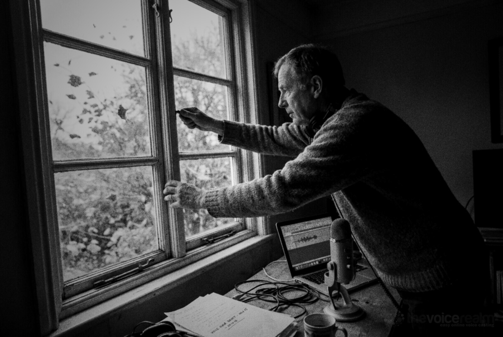 An older man in a wool jumper with headphones round his neck shuts a window in his home recording corner as autumn leaves blow past outside, a microphone and a laptop showing a waveform on the desk beside him, black and white photograph © The Voice Realm