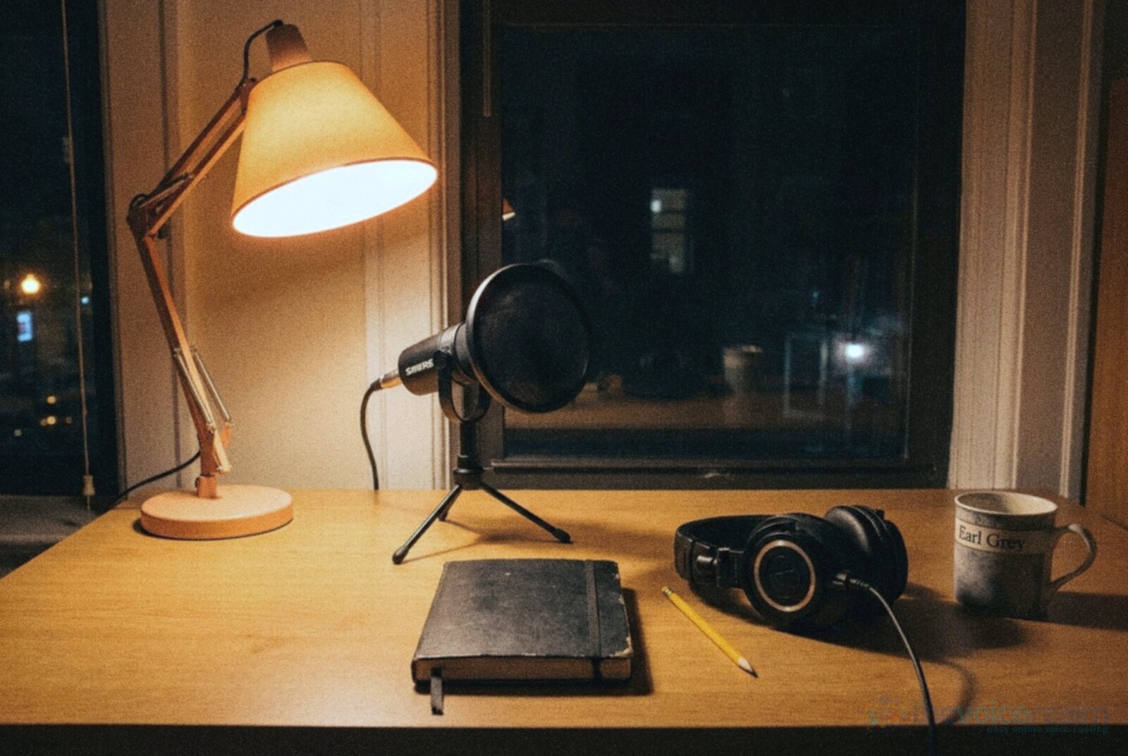 A home voice recording desk at night with a Shure microphone and pop filter on a small tripod stand, closed-back headphones, a closed black notebook, a pencil and an Earl Grey mug under a warm desk lamp, in cool grainy night film colours © The Voice Realm