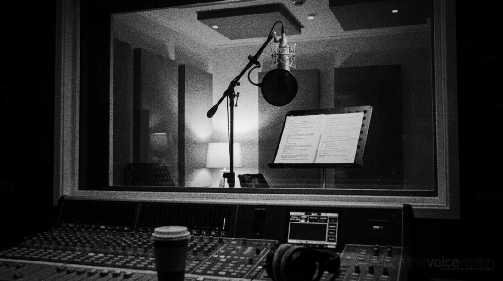 Looking from a dark control room through the glass into an empty voice recording booth with a microphone and pop filter on a stand and a printed script on a music stand, a mixing desk in the foreground, black and white photograph © The Voice Realm