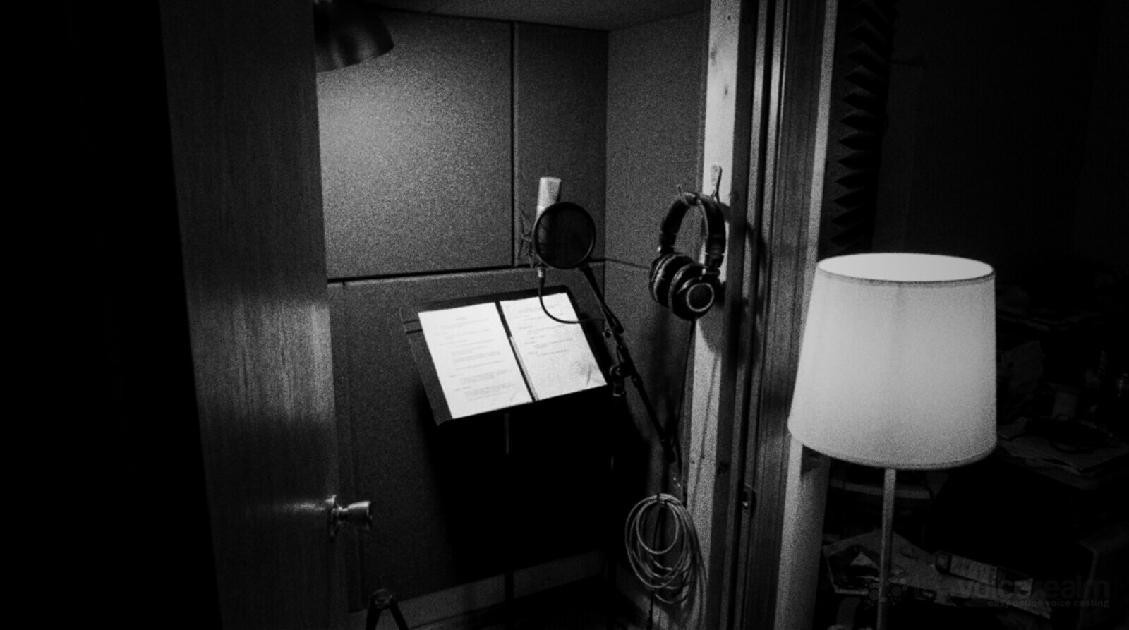 Black and white photo of a small home recording booth at night seen through a half open door, a microphone with a pop filter, script pages on a music stand and headphones on a hook © The Voice Realm
