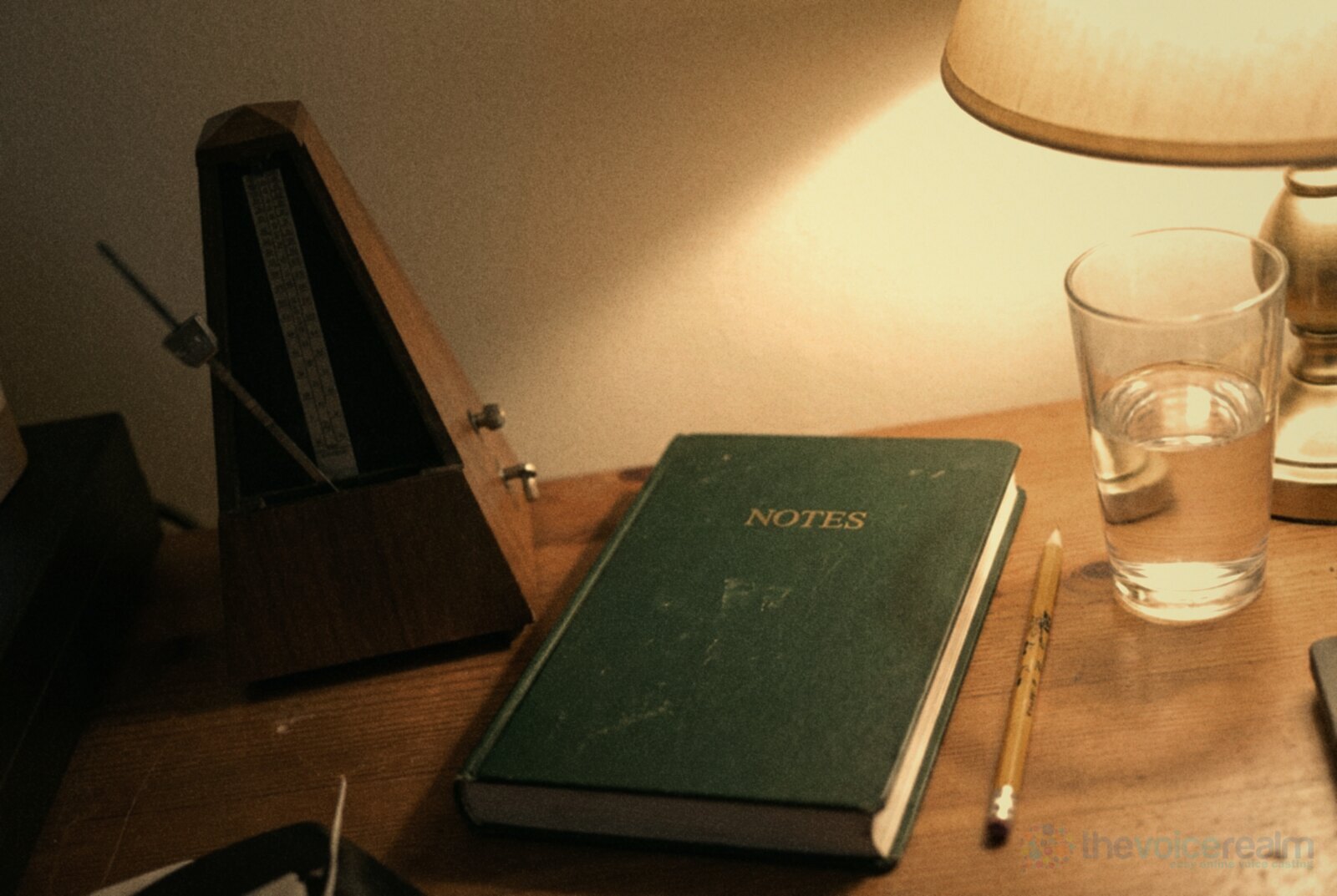 A wooden pyramid metronome with its pendulum mid swing on a desk under a warm lamp, beside a green hardback notebook, a pencil and a glass of water © The Voice Realm