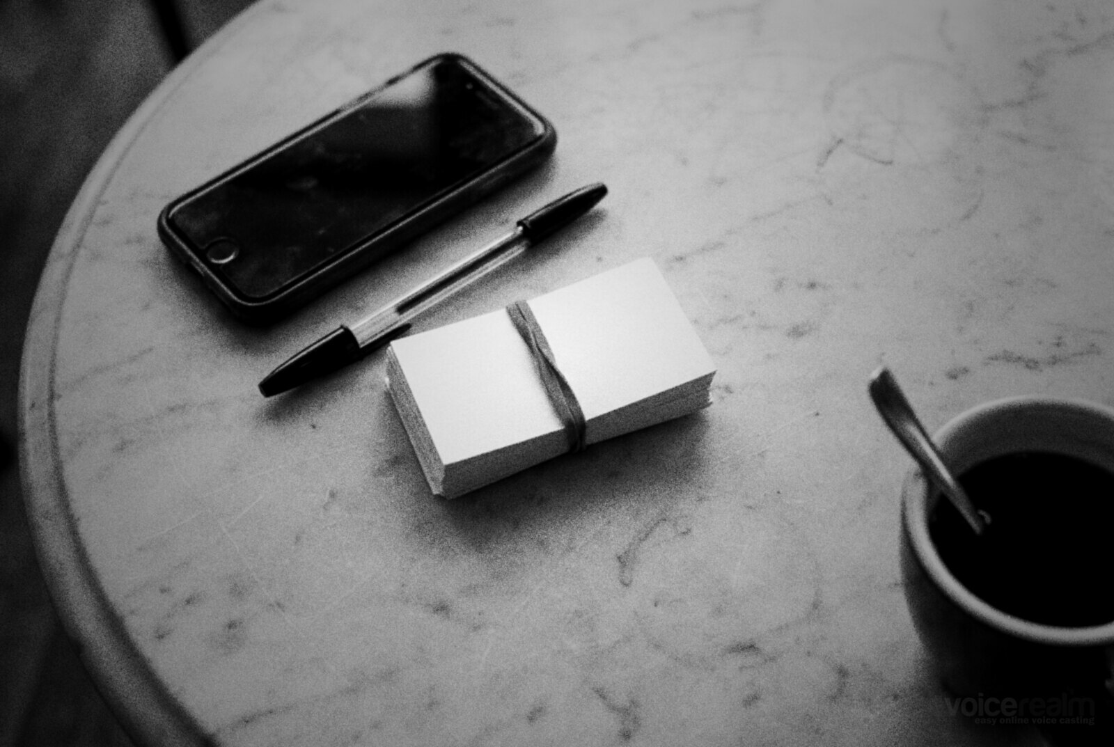 Black and white photo of a stack of blank business cards held with a rubber band, a ballpoint pen and a phone face down on a marble cafe table beside a cup of black coffee © The Voice Realm