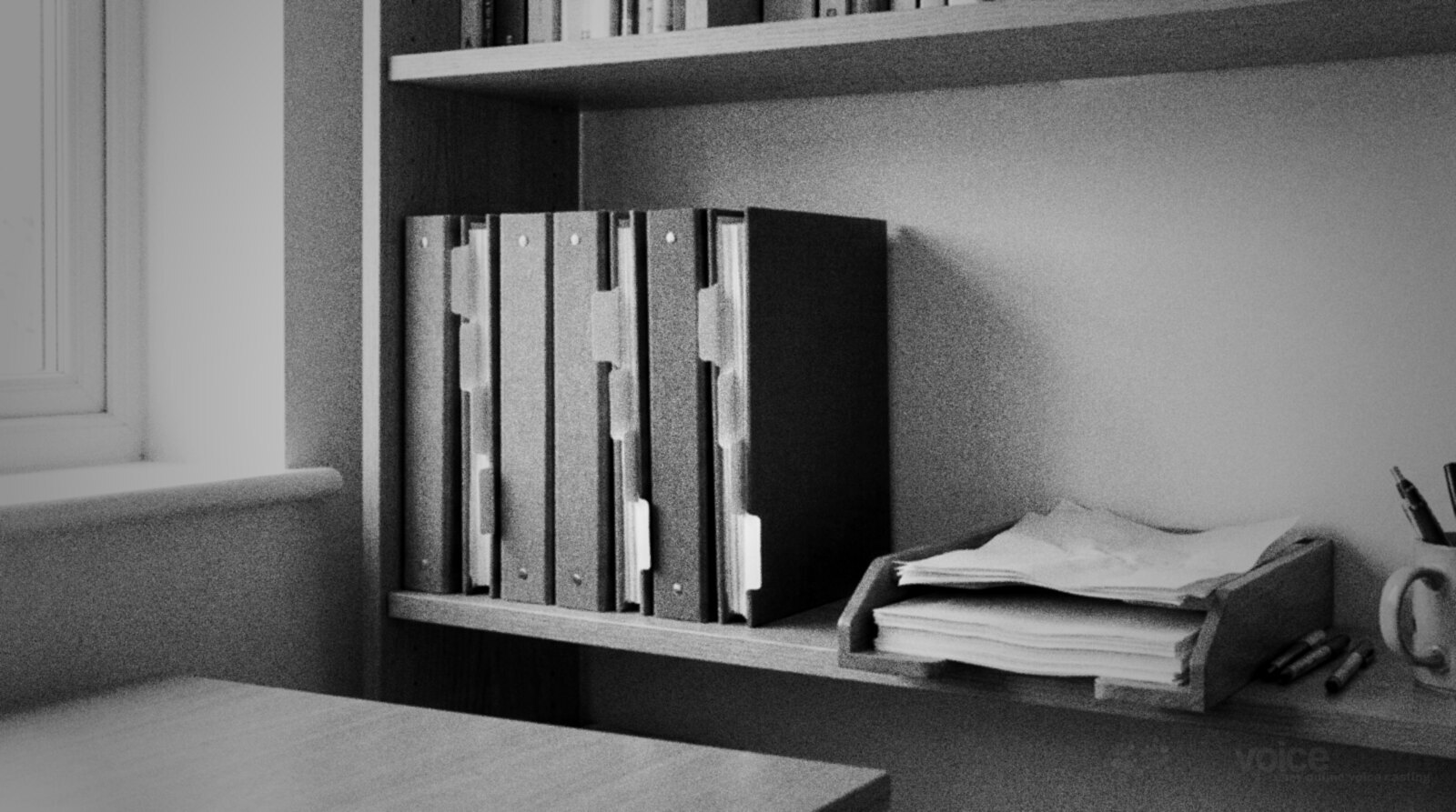 A shelf of tabbed ring binders and a wooden paper tray beside a window in a home office, black and white photograph © The Voice Realm
