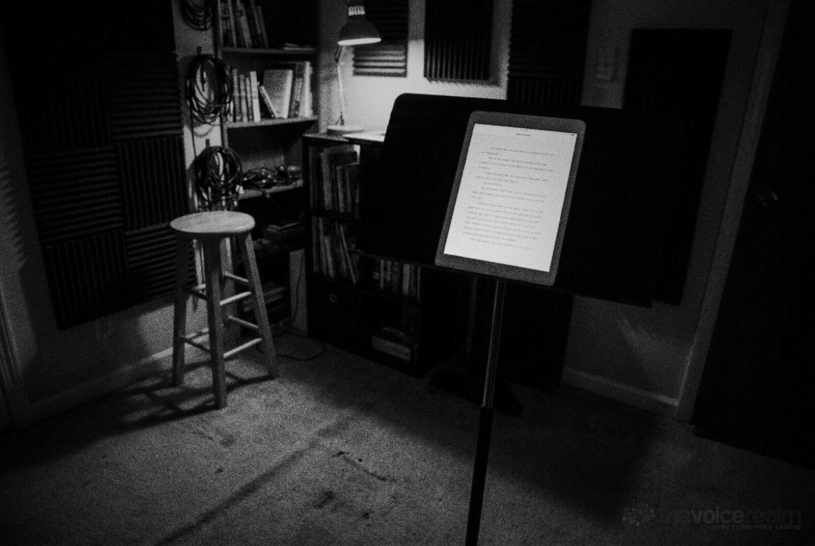 A tablet showing a page of script held at eye height on a floor stand in a small home recording room at night, acoustic foam on the walls, bookshelves, a desk lamp and a wooden stool to one side, black and white photograph © The Voice Realm