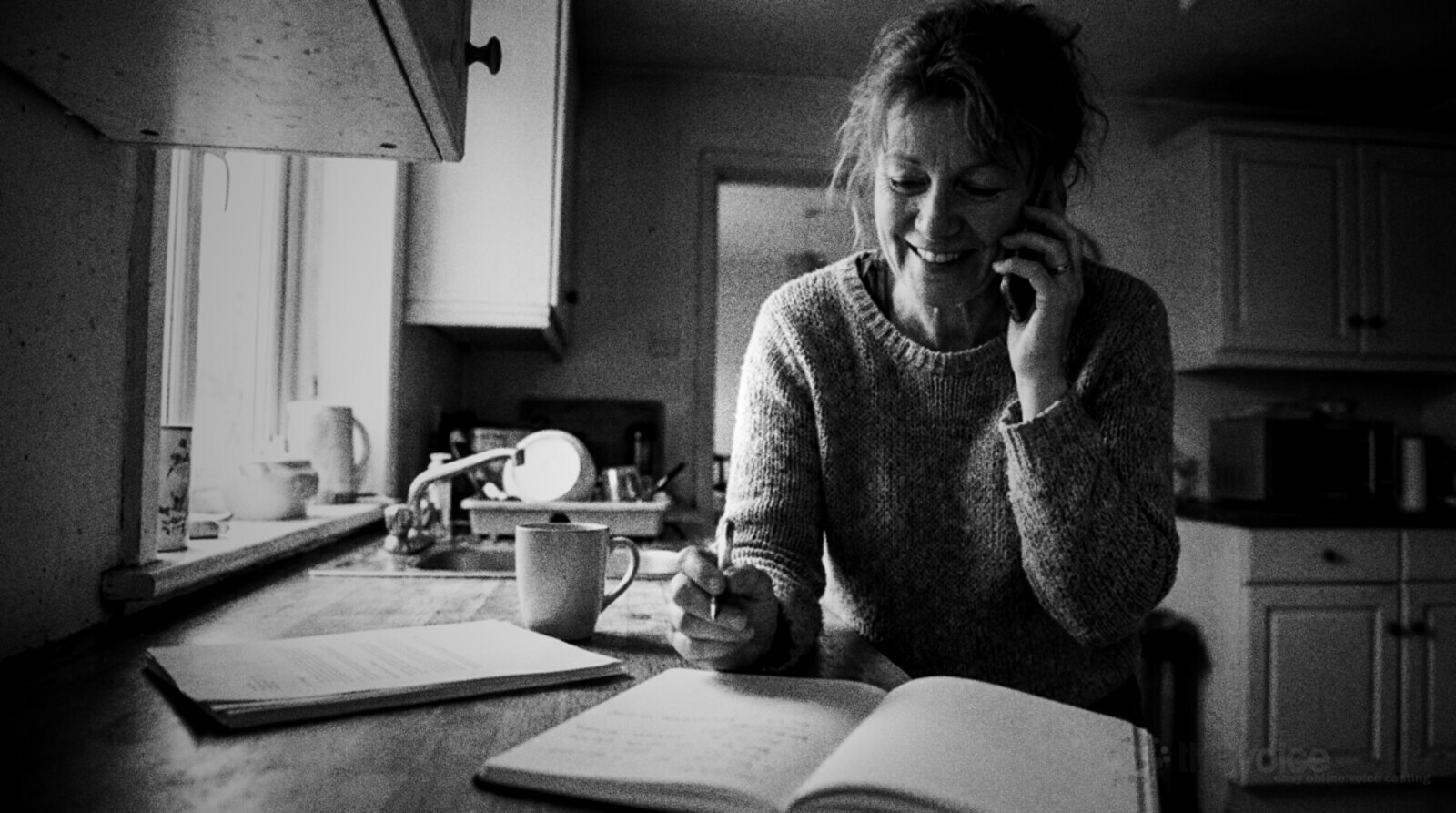 A smiling woman in a knitted jumper on a phone call at a kitchen counter, pen in hand over an open notebook, a mug and a stack of printed pages beside her © The Voice Realm