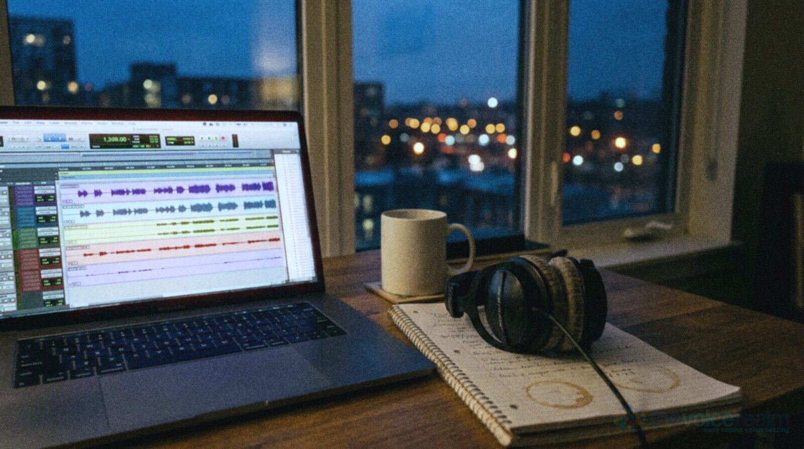 A desk at dusk with a laptop showing several coloured audio waveform tracks, studio headphones resting on a notepad with coffee rings and a plain mug, city lights blurred in the window behind © The Voice Realm