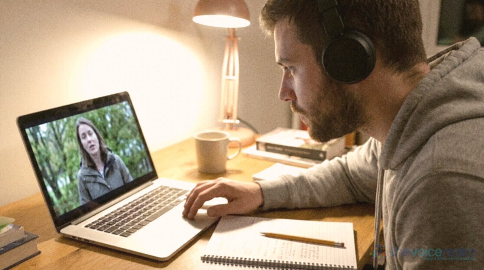 A man in headphones leaning toward a laptop showing a paused interview with a woman outdoors, a notebook and pencil beside him under a desk lamp © The Voice Realm