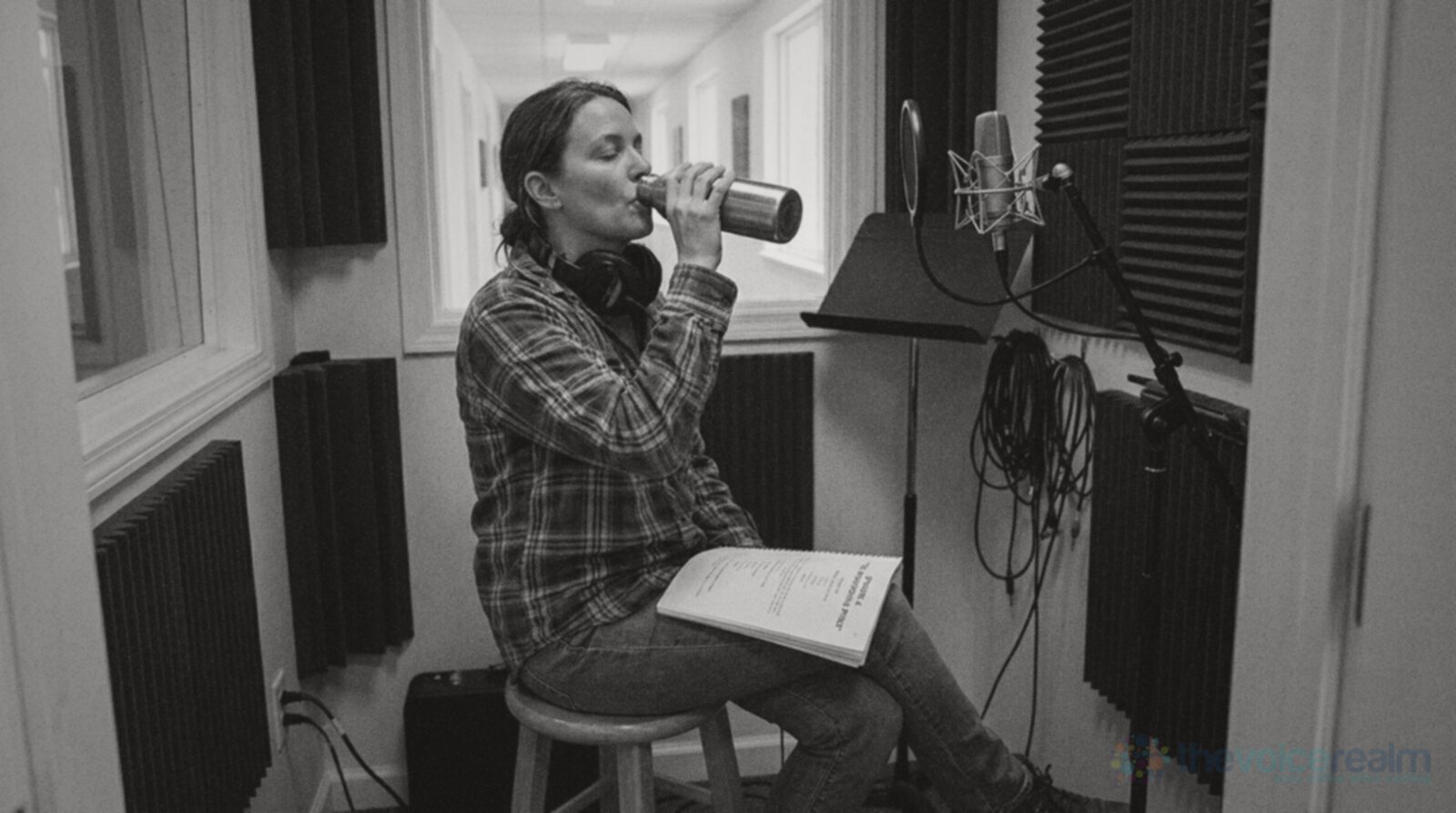 A woman with headphones around her neck sitting on a stool in a recording booth, drinking from a steel water bottle with her eyes closed, a script resting on her knee © The Voice Realm