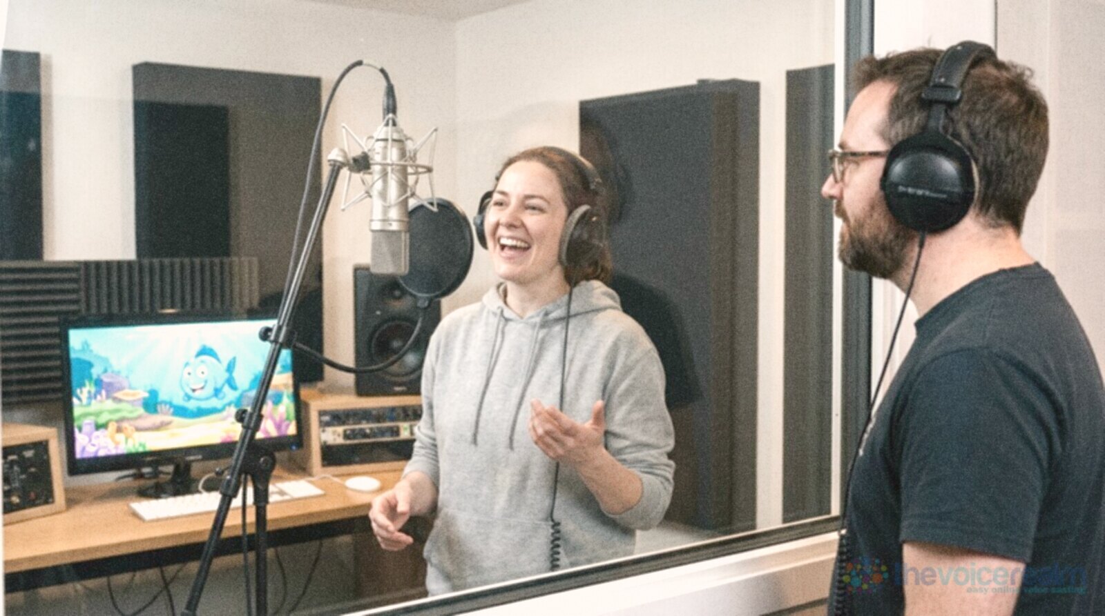 A woman in a hoodie laughing and gesturing at a studio microphone, a colourful cartoon fish on the monitor beside her, a man in headphones watching from the other side of the glass © The Voice Realm