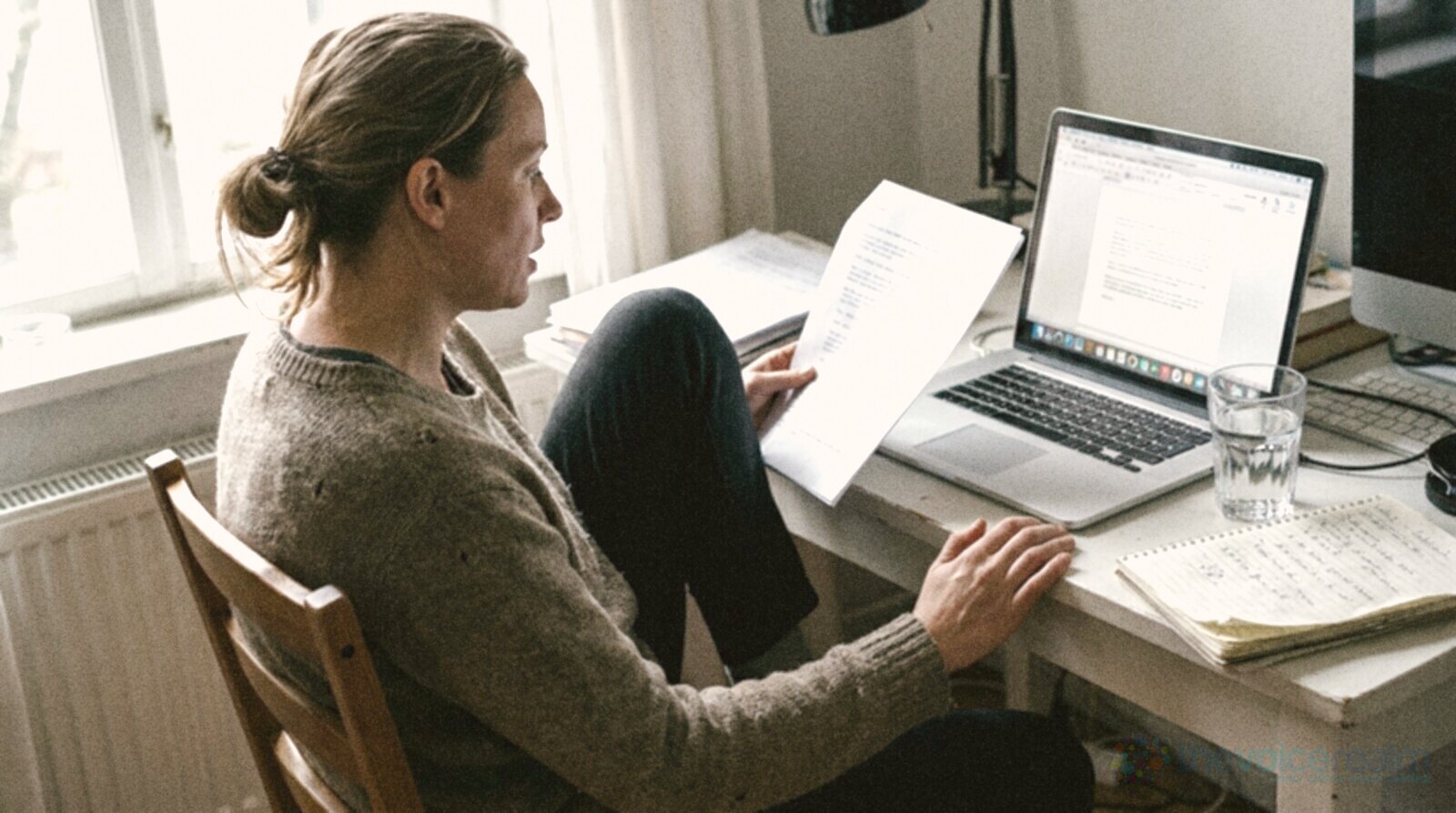A person sitting side-on at a small desk reading a script page aloud in front of a laptop, with a glass of water and a notebook beside them © The Voice Realm