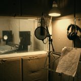 An empty voice over booth lit by a warm lamp: a condenser microphone with a pop filter, headphones on a hook and a marked up script on a music stand, with the control room desk visible through the window © The Voice Realm