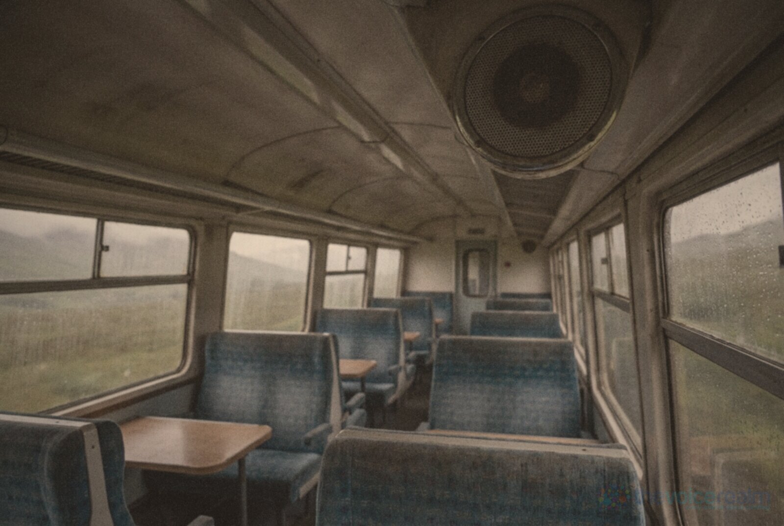 The empty interior of a regional train carriage in Scotland on a rainy day, blue seats, rain on the windows and a round ceiling loudspeaker © The Voice Realm