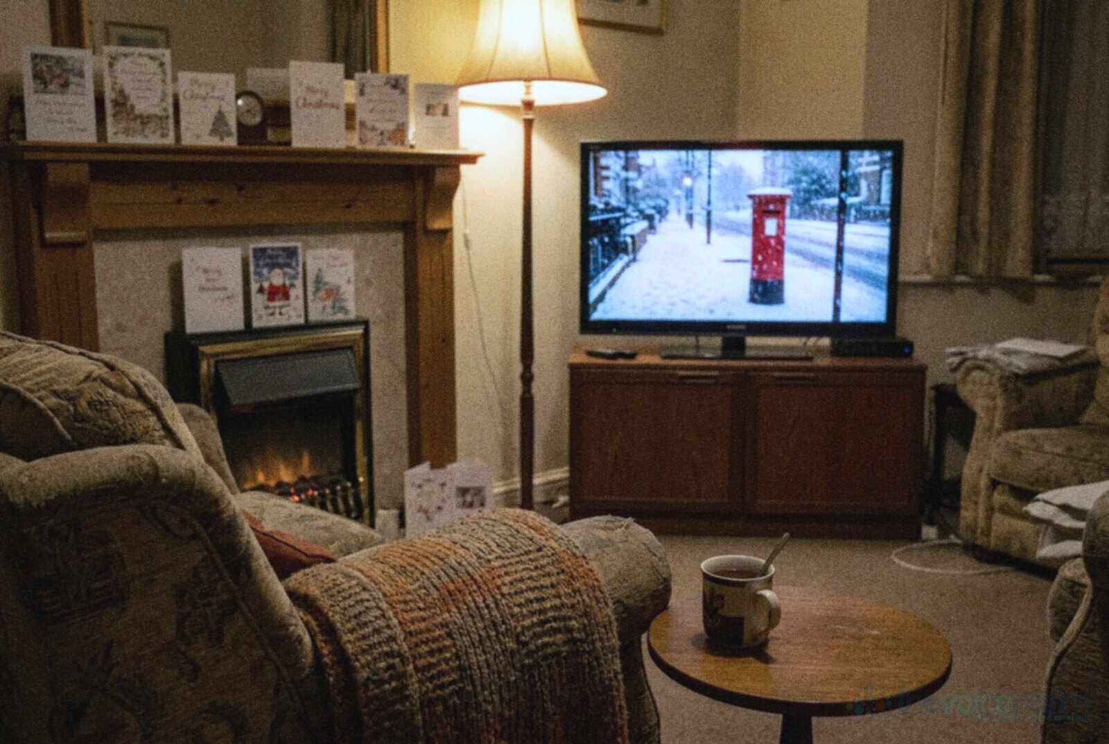 A British living room in the evening with Christmas cards on the mantelpiece, a mug of tea and a television showing a red postbox in falling snow © The Voice Realm