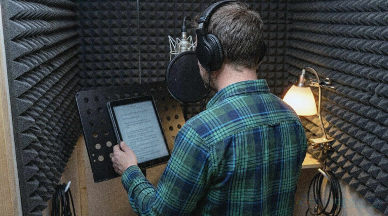A voice actor in a plaid shirt and headphones in a foam lined home booth, reading new copy from a tablet on a music stand in front of the microphone © The Voice Realm