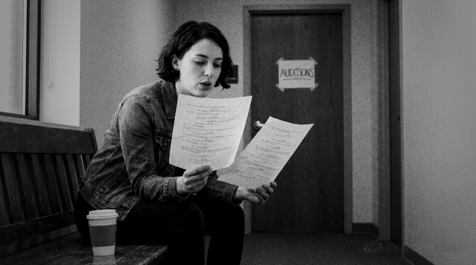 A young voice actor in a denim jacket sitting on a wooden bench in a corridor reading a script aloud for the first time, a coffee cup beside her and a door marked Auditions behind © The Voice Realm