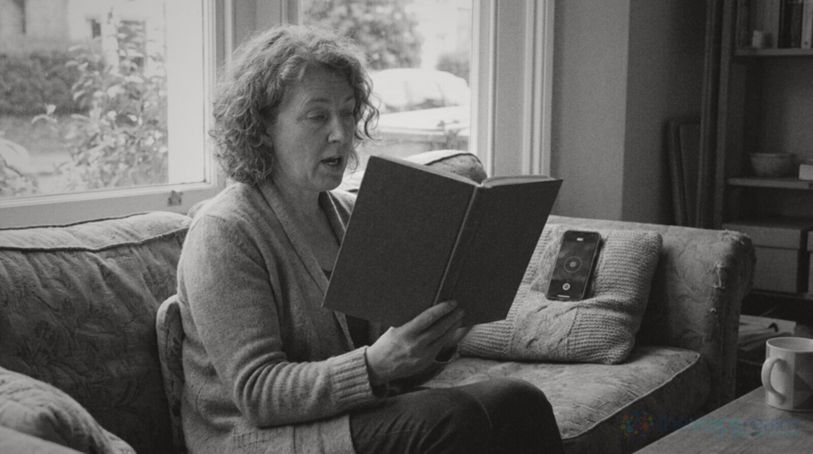 A grey haired woman on a sofa by a window reading a hardback book aloud as practice, her phone recording on a cushion beside her © The Voice Realm