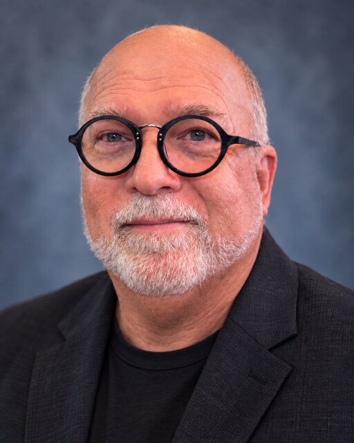 David Atwood, voice actor, in a dark jacket and round black glasses. Photo courtesy of David Atwood