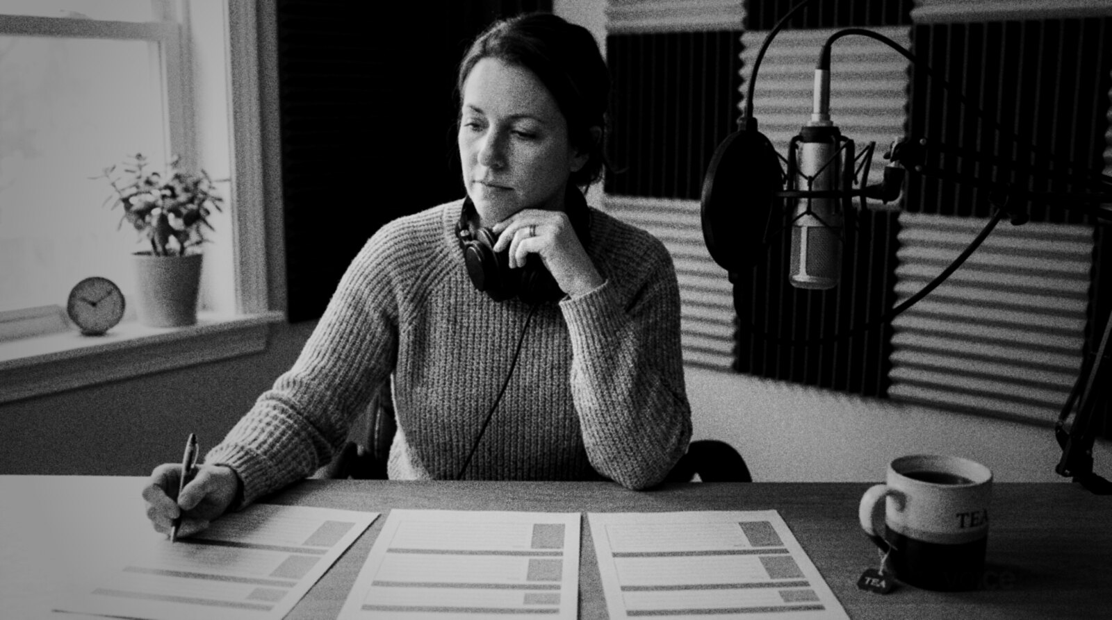 A voice actor at her home studio desk compares three printed sheets laid side by side, pen in hand, with a microphone and acoustic foam behind her © The Voice Realm