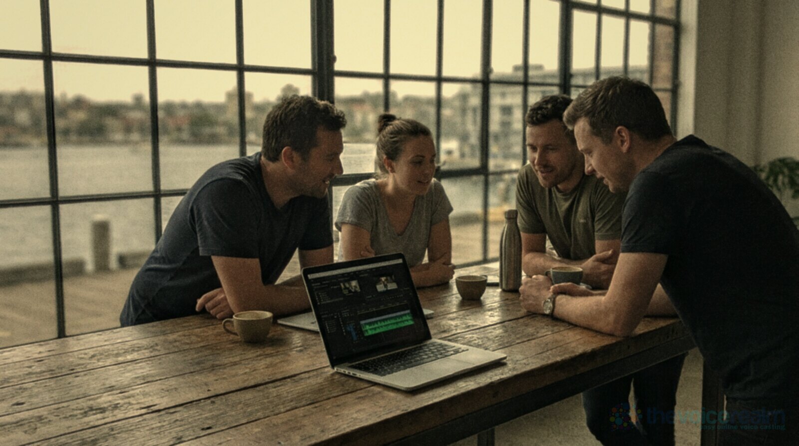 A small Sydney agency team in a converted warehouse by the harbour, gathered around a laptop showing a video edit with an audio waveform © The Voice Realm