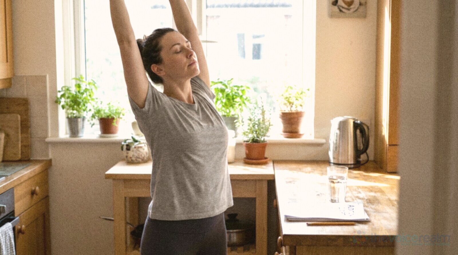 A woman in a grey t-shirt stretching both arms above her head with her eyes closed in a sunny kitchen, a glass of water and a printed audition script with a pencil on the wooden counter beside her © The Voice Realm
