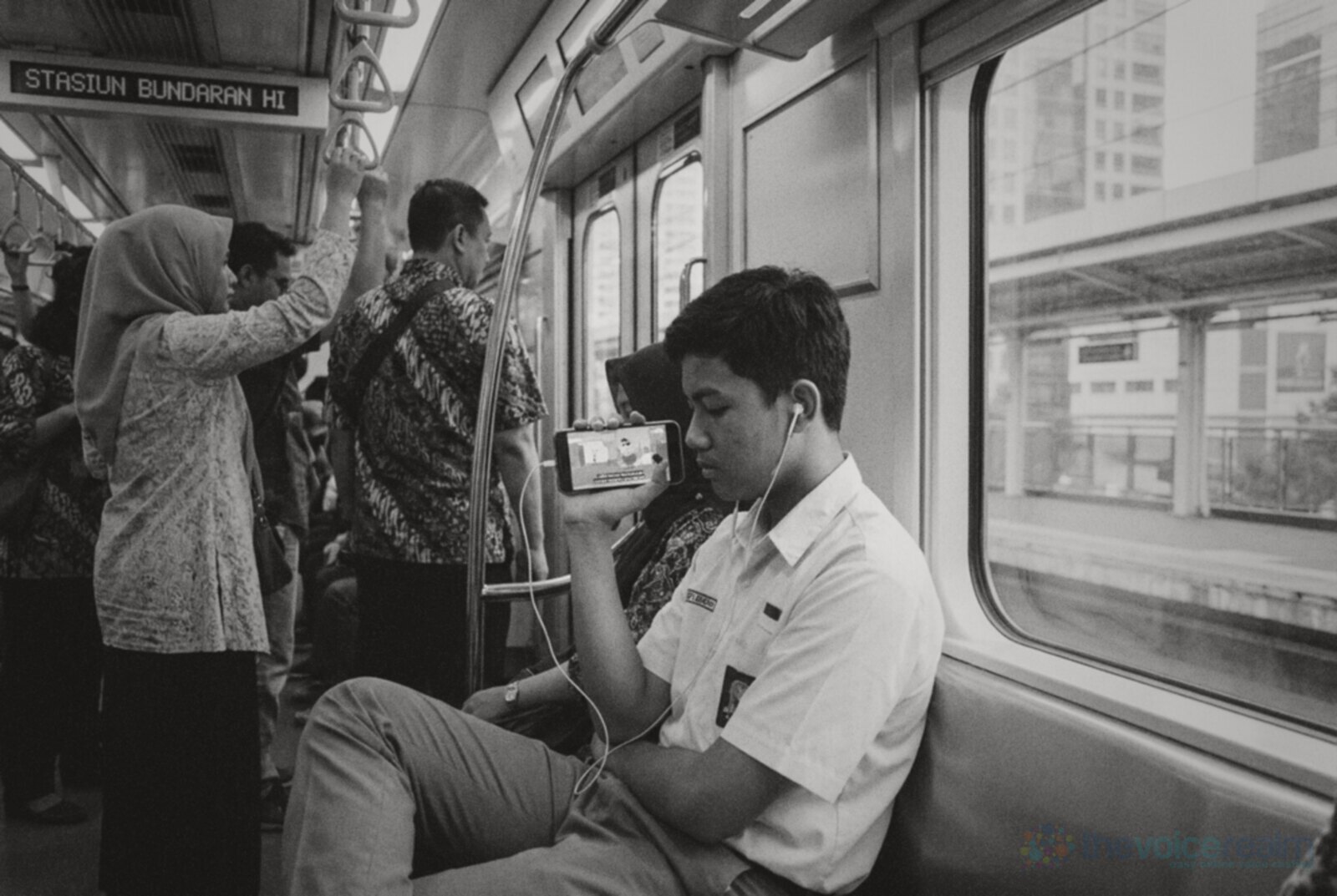 Black and white photo inside a Jakarta MRT carriage: a schoolboy in uniform watches a cartoon on his phone with wired earphones while commuters stand holding straps © The Voice Realm