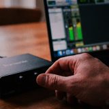 Close up of a hand turning the gain knob on a small desktop audio interface on a wooden kitchen table, a laptop behind it showing a green level meter in a recording program and a mug of coffee beside it © The Voice Realm