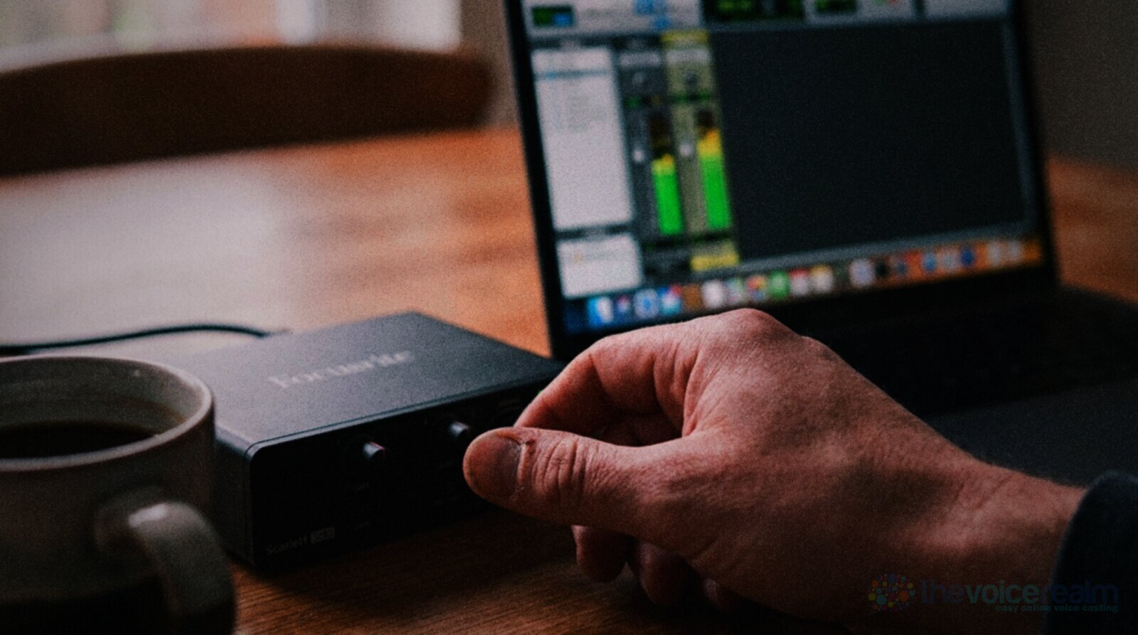Close up of a hand turning the gain knob on a small desktop audio interface on a wooden kitchen table, a laptop behind it showing a green level meter in a recording program and a mug of coffee beside it © The Voice Realm
