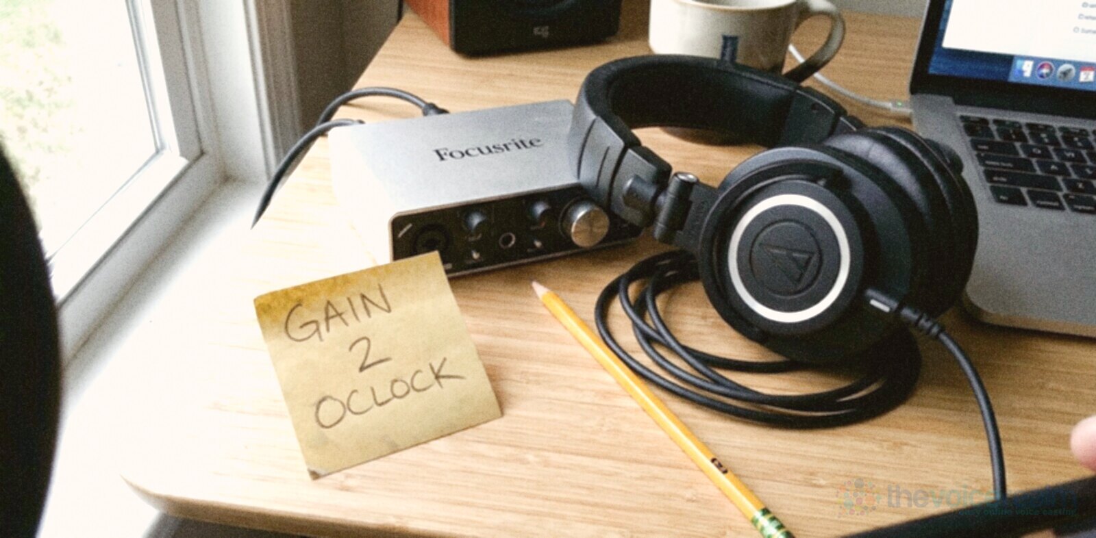 A small silver audio interface and a pair of studio headphones on a home desk by a window, with a yellow sticky note beside them that reads gain 2 o'clock, a pencil and a laptop © The Voice Realm