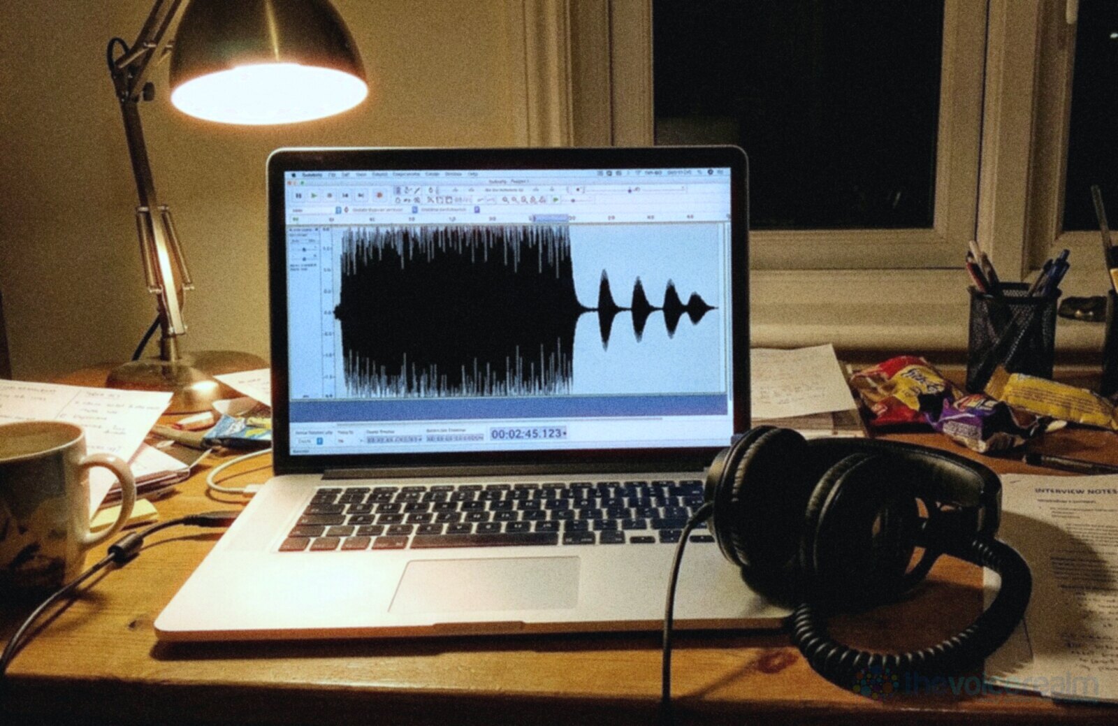 A laptop on a cluttered desk at night under a lamp showing a recording waveform that is tall and flat topped on the left and small with rounded peaks on the right, studio headphones resting beside the keyboard © The Voice Realm