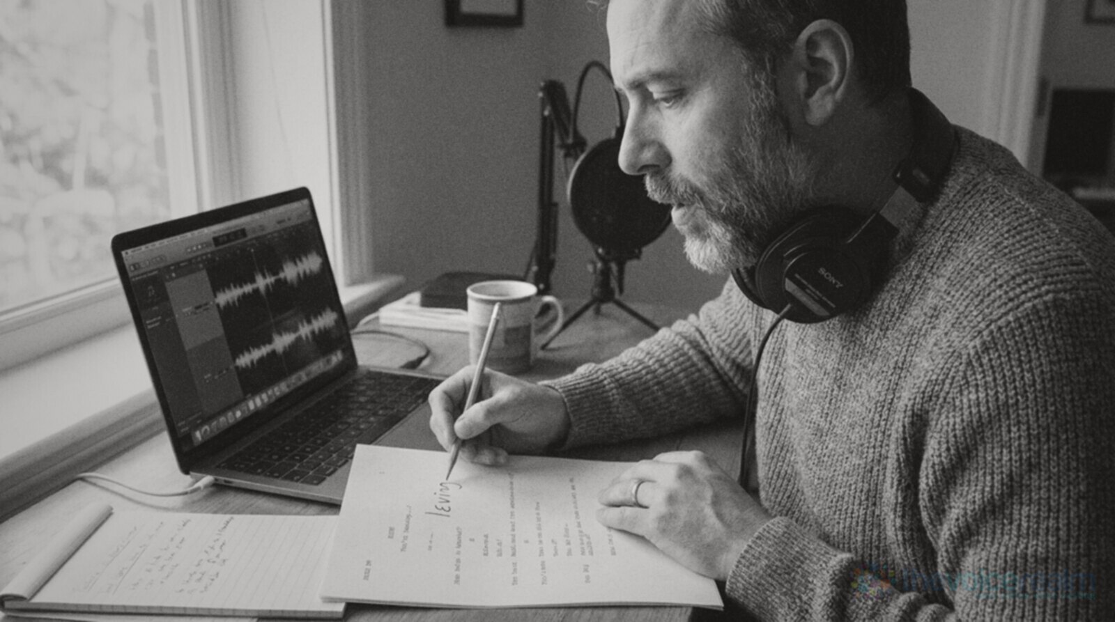 A bearded voice actor in a grey sweater with headphones round his neck at a home desk, saying a word under his breath while pencilling a pronunciation above a line of his printed script © The Voice Realm