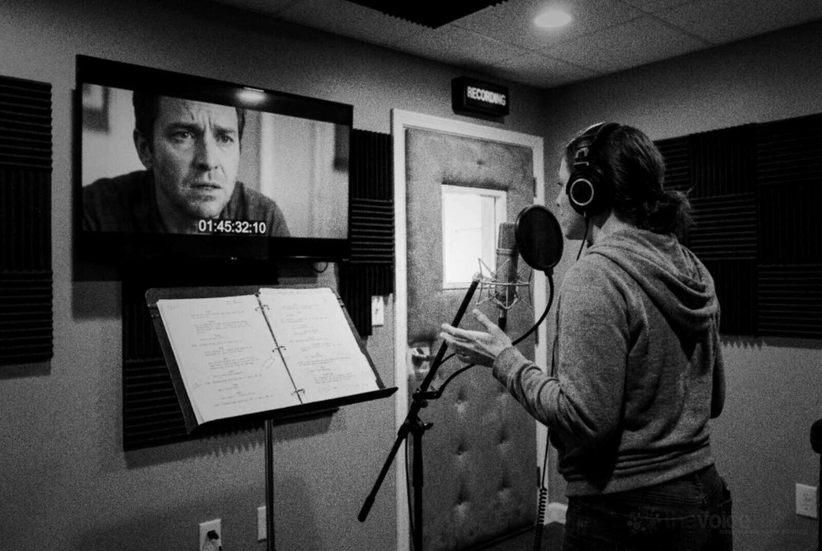 Black and white photo of a voice actor in headphones recording in a dubbing booth, watching an actor's face on a wall screen with a timecode, a script on a music stand beside her © The Voice Realm