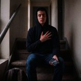 A young man in a black hoodie sitting on the carpeted stairs of an apartment hallway with his eyes closed and a hand on his chest, breathing slowly, a phone in his other hand © The Voice Realm