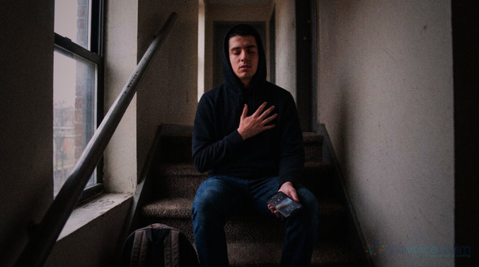 A young man in a black hoodie sitting on the carpeted stairs of an apartment hallway with his eyes closed and a hand on his chest, breathing slowly, a phone in his other hand © The Voice Realm