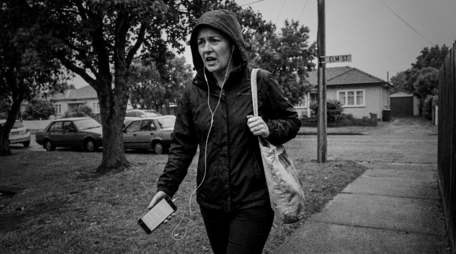 Black and white photo of a woman in a hooded rain jacket walking briskly along a wet suburban footpath with earbuds in, mouthing words to herself, a script on her phone © The Voice Realm