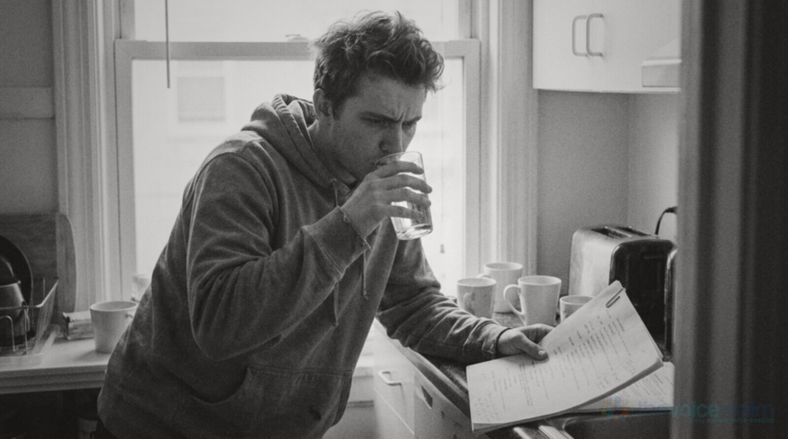 Black and white photo of a young man in a hoodie drinking a glass of water at a small kitchen counter while frowning at a printed script in his other hand © The Voice Realm