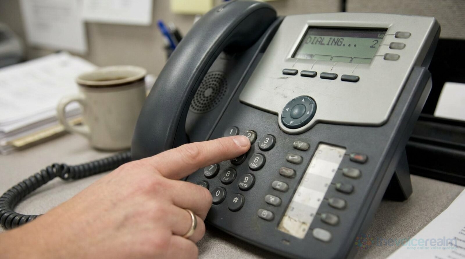 A finger pressing the 2 key on a grey office desk phone, the screen reading Dialing, a coffee mug behind © The Voice Realm