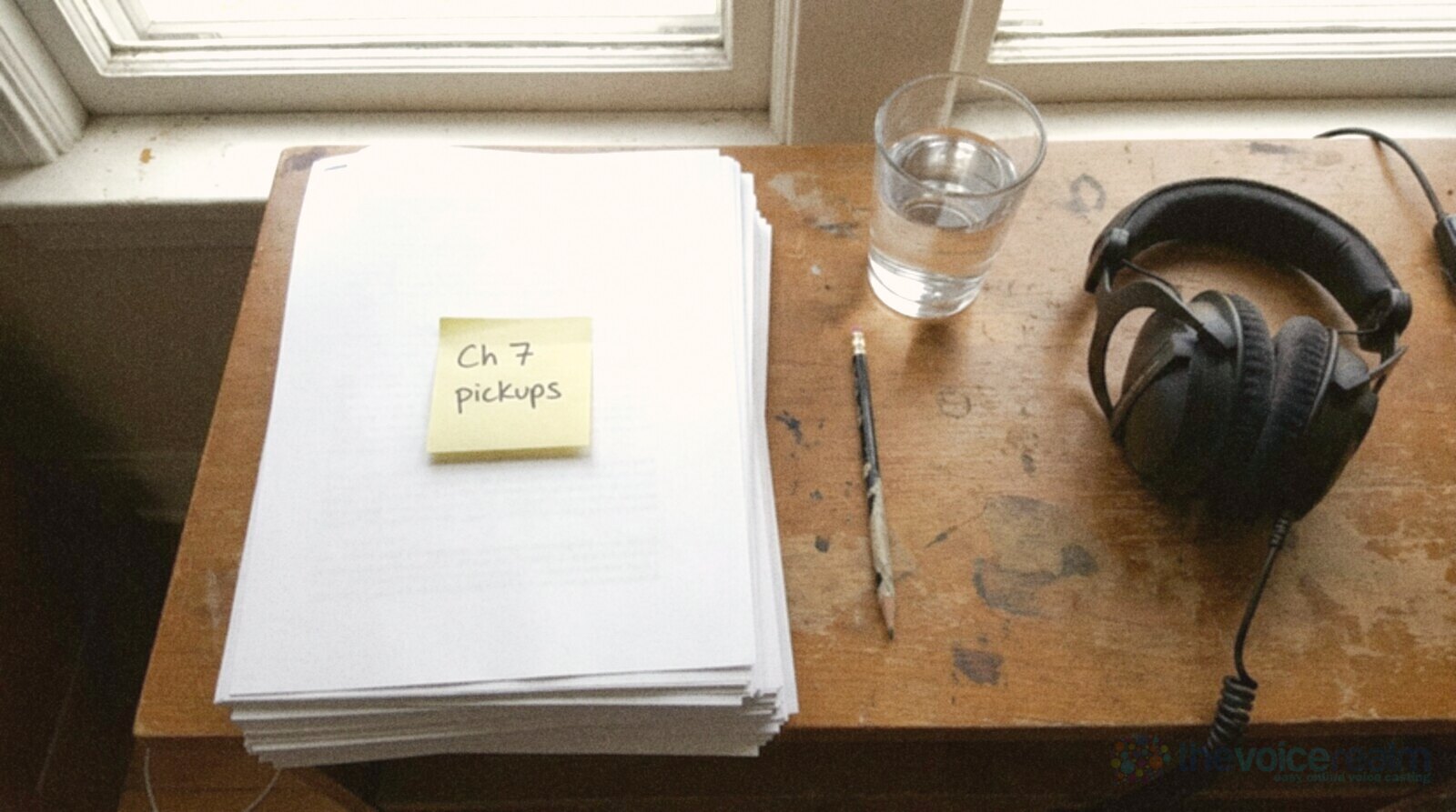 A narrator's wooden desk by a window with a stack of printed pages face down, a yellow sticky note reading Ch 7 pickups, a pencil, a glass of water and studio headphones © The Voice Realm