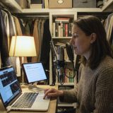 A narrator in a knitted jumper recording in a converted walk-in wardrobe at home, coats hanging behind her, a condenser microphone in front of her, a tablet showing a chapter of a book and her finger on the laptop keyboard © The Voice Realm