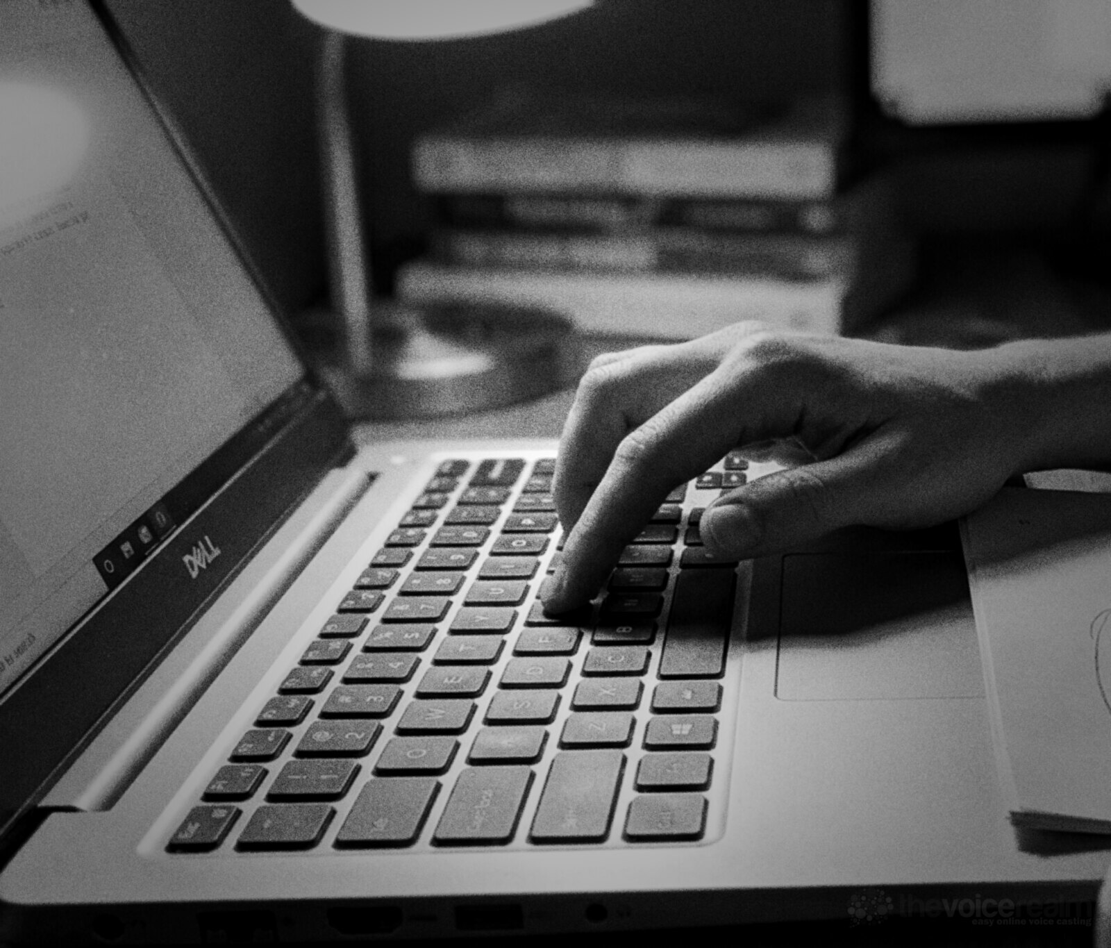 Black and white close up of a hand pressing a key on a laptop keyboard at a desk at night, a lamp and a stack of books behind © The Voice Realm