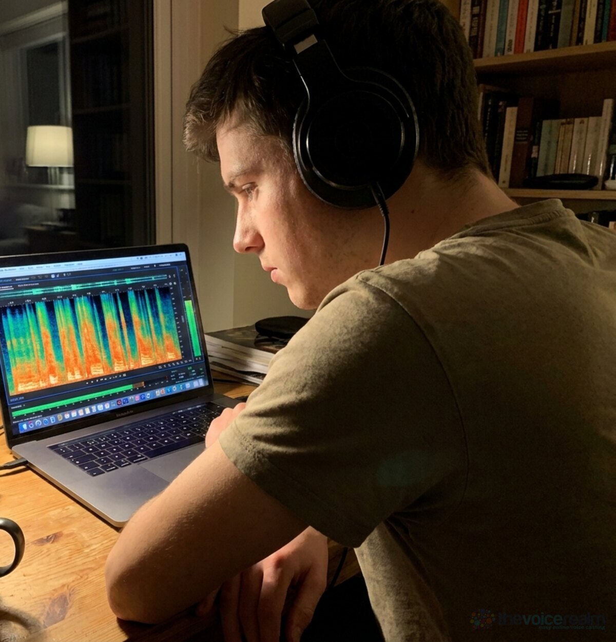 A young voice actor in headphones at a desk at night studying a spectrogram of his recording on a laptop, bright streaks at the top of the display marking each S © The Voice Realm