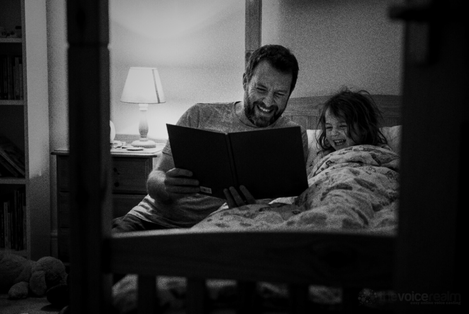 Black and white photo of a father with a beard reading a bedtime story aloud with a big grin while his young daughter laughs under the covers, seen through the bars of the bed frame © The Voice Realm