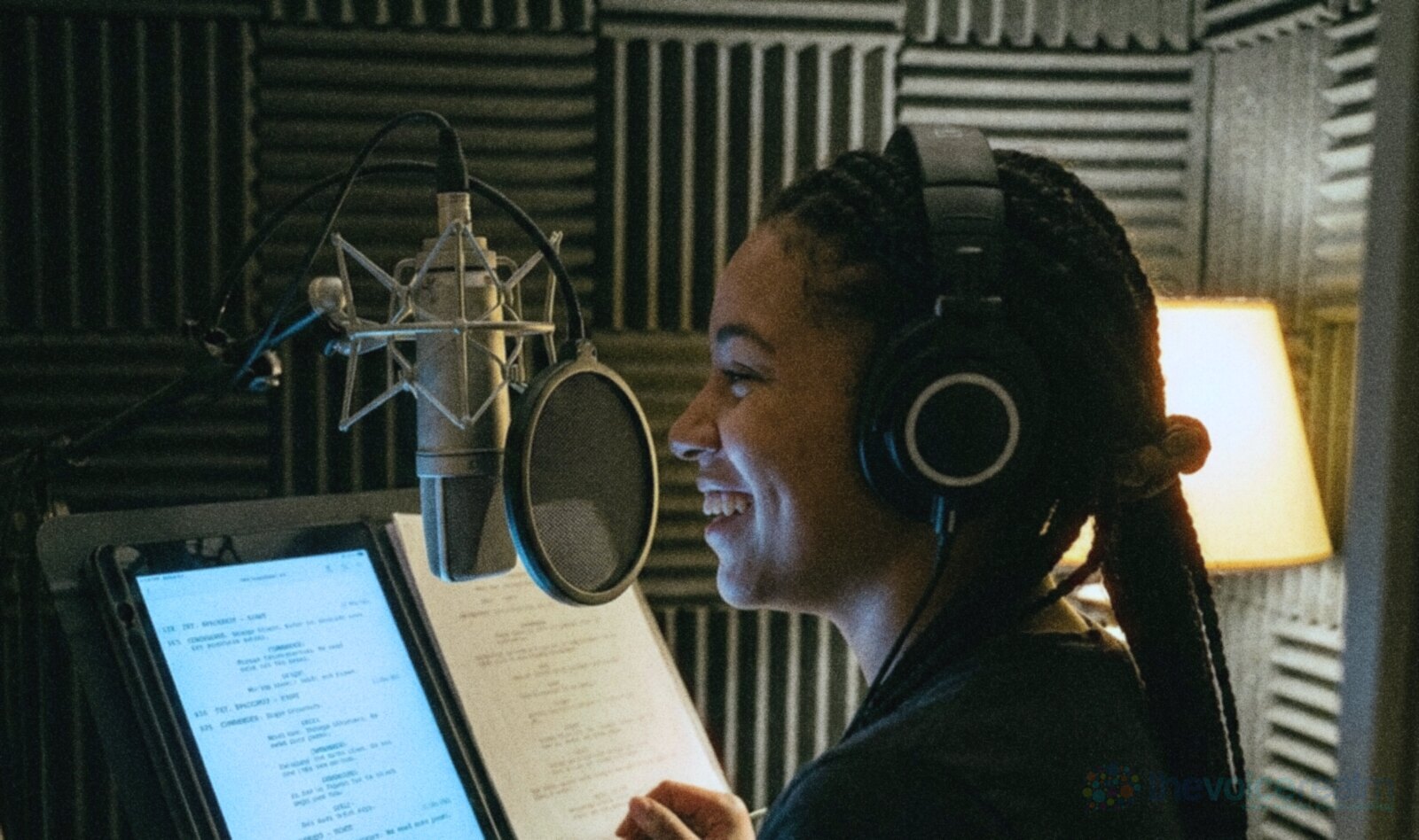 A young voice actor with long braids and headphones grinning as she reads a script from a tablet in a small foam-lined home booth at night, a condenser microphone and pop filter in front of her © The Voice Realm