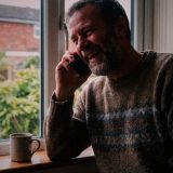 A man in his forties in a knitted jumper laughing warmly on a phone call at his kitchen window, a mug on the window sill and a garden behind him © The Voice Realm