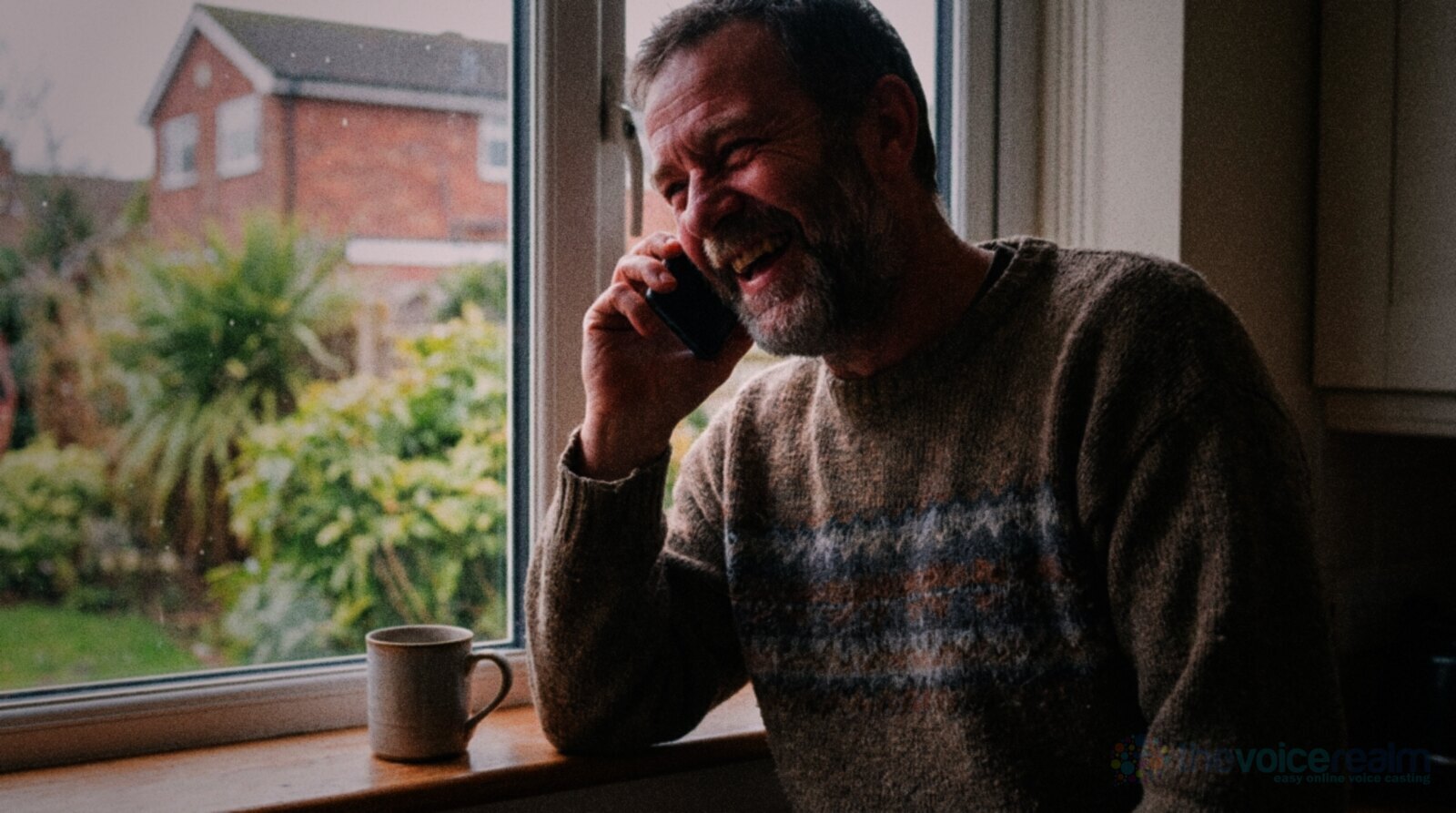 A man in his forties in a knitted jumper laughing warmly on a phone call at his kitchen window, a mug on the window sill and a garden behind him © The Voice Realm